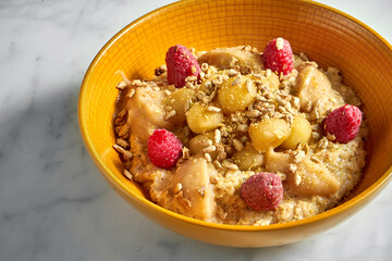 Tasty and healthy oatmeal with honey, apple, raspberry and sunflower seeds in a bright yellow bowl on a white marble background.