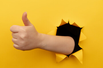 Female hand showing a thumb up sign through a ripped hole in yellow paper wall. Well done, good job...