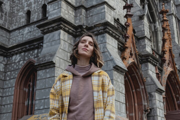 Happy millenial woman with closed eyes enjoys in a yellow coat in European city near historic building. Local travel concept.