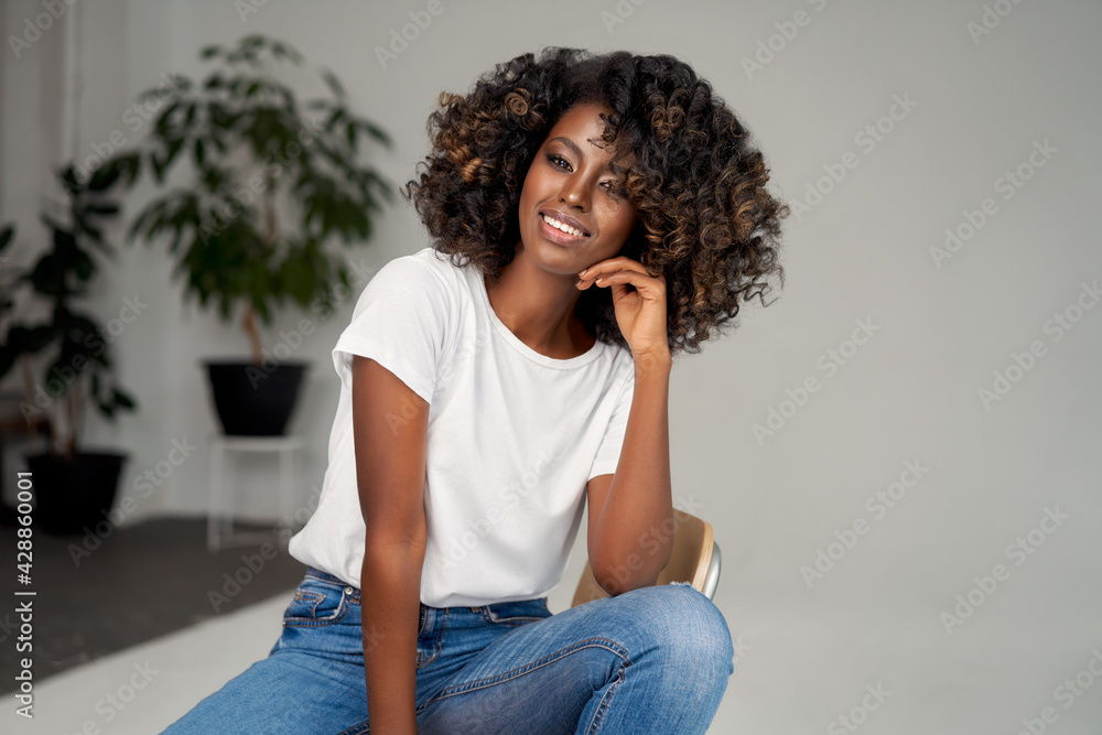 Wall mural Beautiful black woman sitting on chair. Cheerful african american woman relaxing at home.