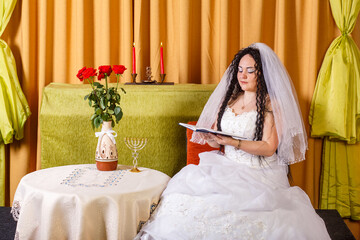 A Jewish bride in a white wedding dress with a veil sits at a table with flowers and reads blessings from a prayer book before the chuppah ceremony.