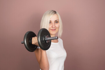 Beautiful young blonde woman with big dumbbells is engaged in fitness on a pink background