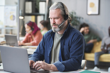 Mature bearded man in headphones working on laptop at the table in call center