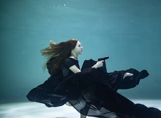 beautiful girl in a black swimsuit with a black pareo holds a black pistol in her hands on a blue background underwater