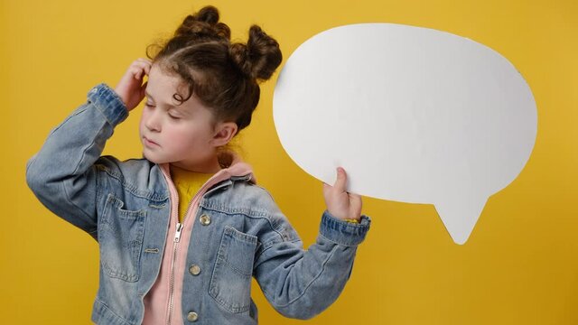 Thoughtful little girl holding an empty white speech bubble, ponders on decision, thinking about something, wears denim jacket, isolated over yellow background with copy space. Hmm something strange