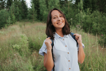 A girl-traveler stands against the background of the forest with a backpack and looks into the...