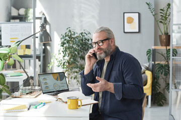 Mature businessman in eyeglasses sitting at his workplace and talking with his colleague on mobile...