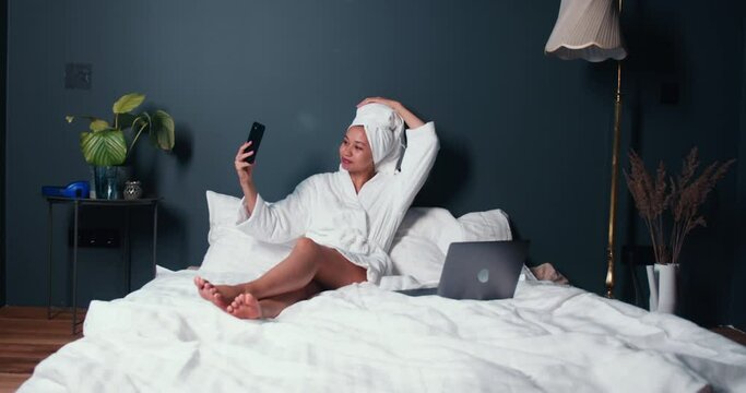 Young happy successful mixed race blogger woman in white bathrobe takes smart phone selfie picture sitting on large bed.