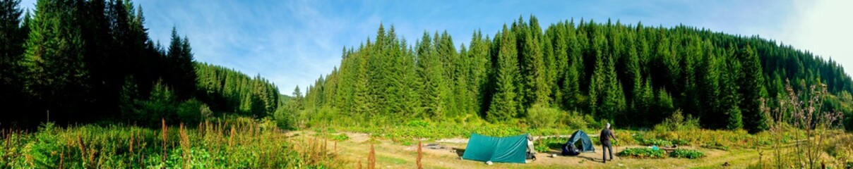 Panorama of a tourist tent on a green meadow on a background of forests and peaks