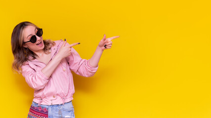 A young caucasian excited smiling  woman in a pink jacket and sunglasses indicate copy empty blank space for text or design with two fingers presenting product on bright color yellow background.Banner
