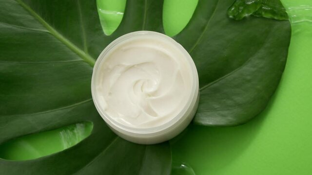 Cosmetic jar with cream on green tropical leaf in clean water splash on green background. Top view. Plant-based skincare, organic natural ingredients, skin routines, summer cosmetics, fresh concept