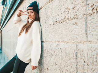 Portrait of beautiful smiling model. Female dressed in warm hipster white sweater and beanie. Posing in the street at sunset. Funny and positive woman having fun outdoors