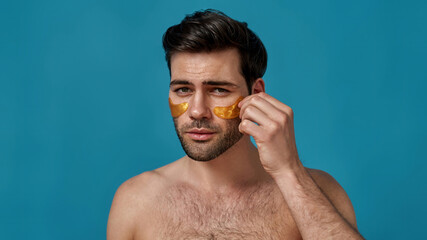 Good looking brunette guy applying golden gel under eye patches on his skin while looking at camera, posing isolated over blue background