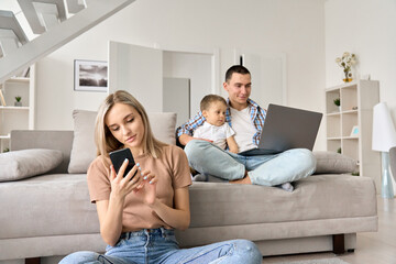 Young family at home, dad with kid sitting on couch watching online video early educational classes, movies on laptop, mother holding cell phone surfing social media, spending leisure time at weekend.