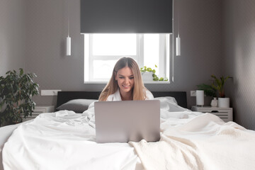 Smiling girl lays on the bed in bedroom and use laptop for remote work
