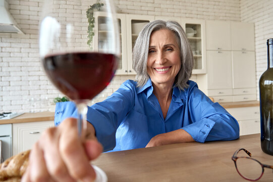 Senior Mature 60s Aged Older Woman Sitting At Kitchen Home At Table With Computer. Having Virtual Wine Party With Friends Family On Remote Video Call Meeting Clinking Glass To Camera, Web Cam View.