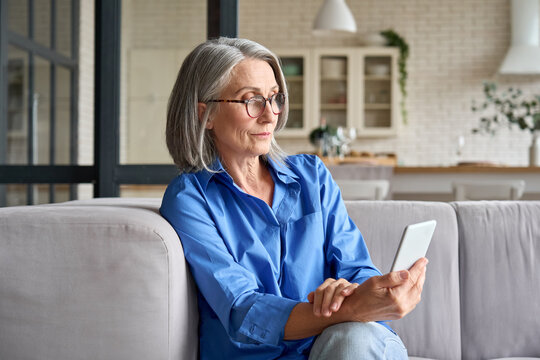 Serious mature middle age senior woman at home on couch holding mobile cellphone, reading news or watching online learning class having video call using mobile application.
