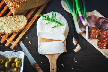 Mozzarella cheese sliced on a wooden plate with freshly baked bread with green onions, smoked ham, sausage, olives on a dark wooden dining table.Top view
