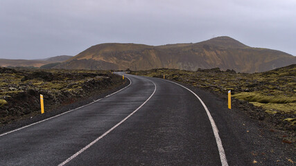 Fototapeta na wymiar Diminishing perspective of winding country road with markings between moss covered lava fields of volcanic stones near Grindavik, Reykjanes peninsulsa, Iceland with Þorbjörn mountain in background.
