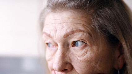 Close up on blue sad eyes of lonely elderly woman looking through the window. High quality photo