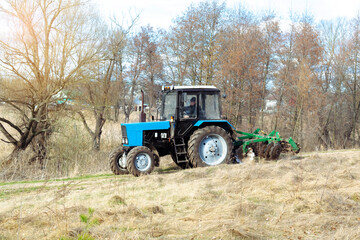 Blue small modern tractor machinery plowing agricultural field meadow at farm on sunny day. Farmer cultivating and make soil tillage before seeding plants and crops, nature countryside rural scene.