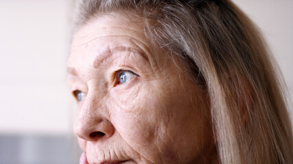 Close up on blue sad eyes of lonely elderly woman looking through the window. High quality photo