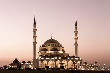Naklejka premium New Sharjah Mosque, the largest mosque in the Emirate of Sharjah, the United Arab Emirates, with pink sky sunset and facade illuminations in the evening.