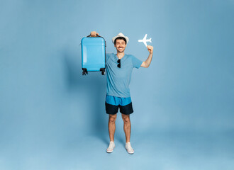 Full-length photo of a happy excited caucasian guy in a hat holds a blue suitcase and a small...