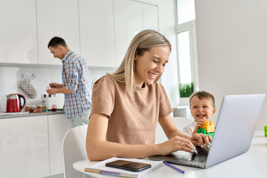 Happy Young Mom Business Woman Distance Working Online Using Laptop Computer While Small Funny Kid Son Playing Having Fun Sit At Kitchen Table. Virtual Homeoffice Work And Child Care In Family Life.