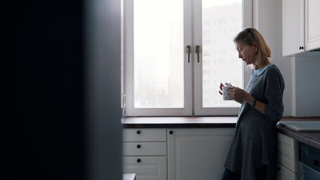 Lonely elderly woman drinking coffee standing at her kitchen. High quality photo