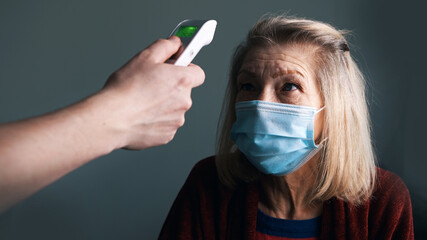 Digital thermometer measuring body temperature of an elderly woman with medical mask. High quality...