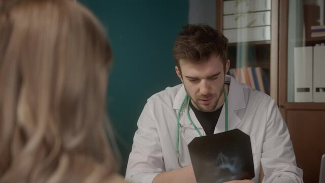 Confident young American doctor reading x-ray picture, concentrated facial expression, treating patient online using video chat internet connection on laptop computer