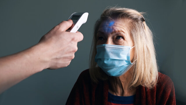 Check up of body temperature with digital thermometer. Old gray haired woman with face mask having dotor visit at home. High quality photo