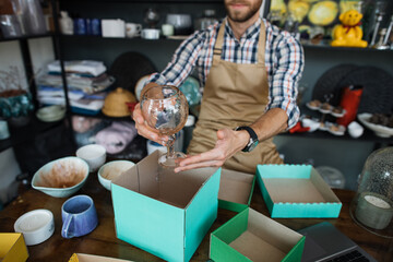 Close up of professional seller in apron doing inventory at decor store. Young man sitting at counter and checking goods for no damages.