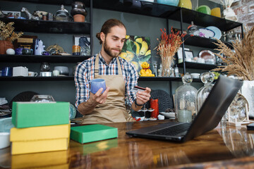 Bearded seller in beige apron preparing discount card for regular customer at decor store. Handsome man sitting at counter and working on modern laptop.