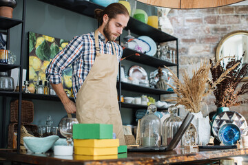 Handsome caucasian man wearing beige apron working on modern laptop at decor store. Qualified seller creating online catalogue with available goods at shop.