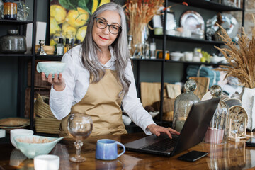 Attractive mature woman standing at counter with laptop and various decor, smiling and looking at camera. Inventory process at modern store.