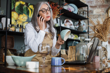 Professional saleswoman with grey hair sitting at decor shop with opened laptop and talking on mobile. Aged woman using modern gadgets at work.