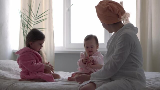 Authentic cute young woman combing long hair of baby girl. mom with 2 children daughters different ages in bathrobes after bath on white bed oposite window. Spa, motherhood, childhood, family concept.