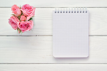 A checkered notebook and pink roses on a white wooden table. The scheduler. Empty space. Massage Flatt lay. Top view.