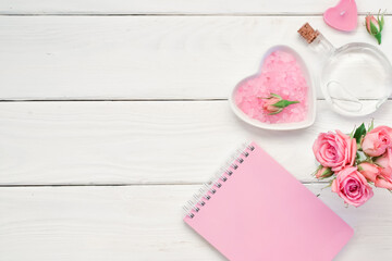 A notepad and pink roses on a white wooden table with space for text. Bath salts and perfumes. Empty space. Flatt lay. Top view.