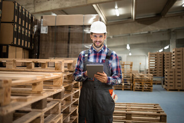 Portrait of warehouse worker typing on tablet computer and standing by wooden palette in factory storage room.