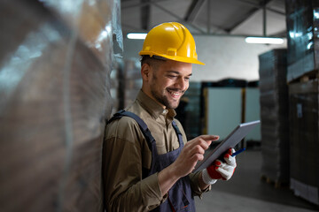 Portrait of caucasian smiling warehouse worker standing by palettes and checking inventory on tablet computer in storehouse.