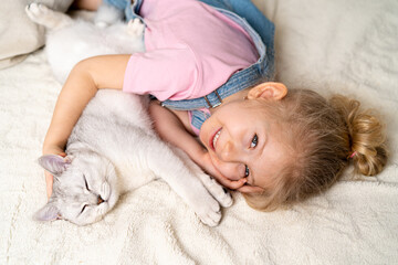 Little blonde girl lies with a white kitten on a soft blanket smiling and looking into the camera