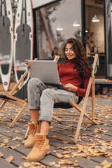Beautiful woman in cozy outfit works at laptop while sitting on chair in autumn park.