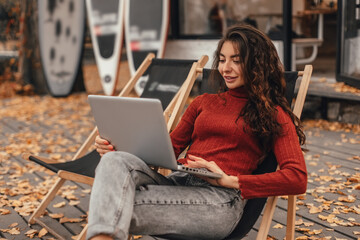 Beautiful woman in cozy outfit works at laptop while sitting on chair in autumn park.