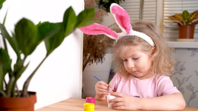 a little blonde girl is decorating an Easter egg and smiling, at home at the table. the concept of a religious holiday