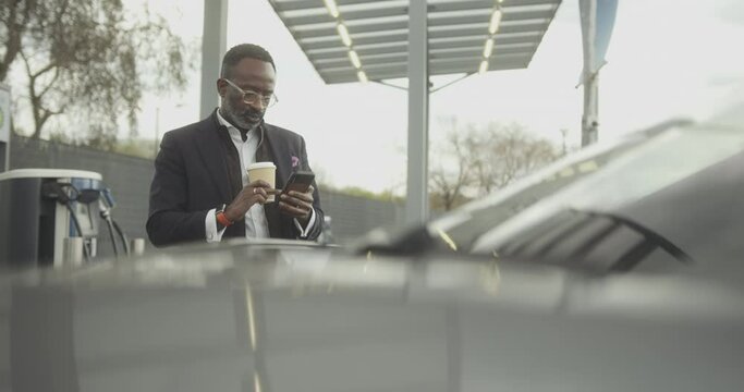 Business man charging electric car at charging station looking at smart phone and drinking coffee
