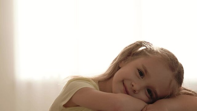 Portrait of super cute little girl with two ponytails looking at camera and smiling while resting at home, selective focus on lovely child lying on table. Kids spending leisure time indoors