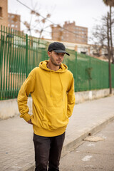 Young caucasian male in a yellow hoodie and black cap standing on the street